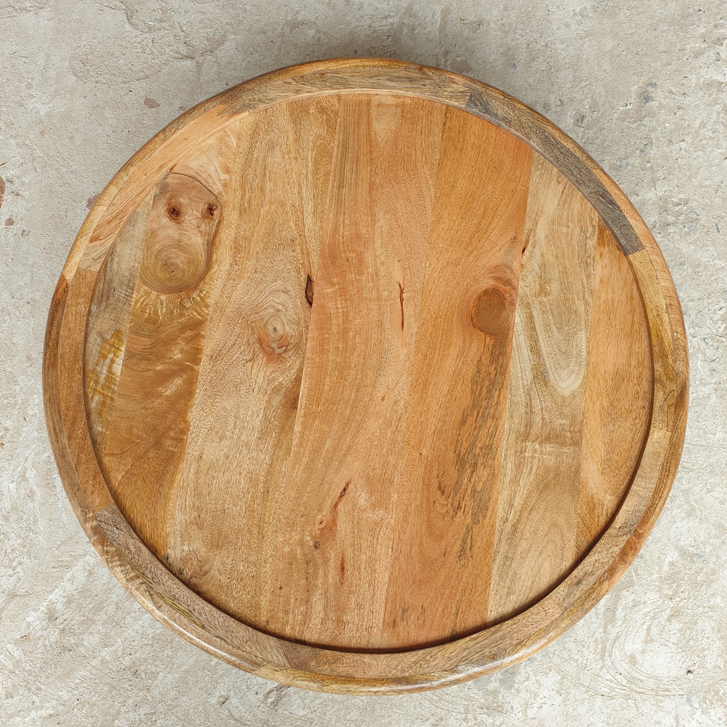 A round wooden serving board or tray with a raised rim, made from natural wood with visible grain, knots, and warm tan and brown tones, photographed from directly above on a light gray textured surface.