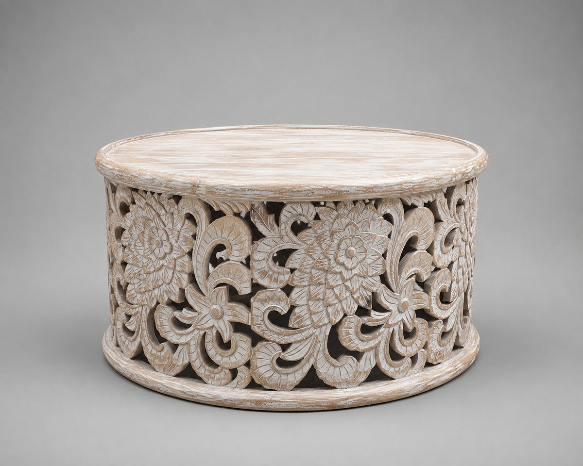 A round carved wooden stand or low table with an intricate floral and scroll cutout pattern, finished in a distressed off-white finish, set against a solid gray background.