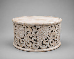 A round carved wooden stand or low table with an intricate floral and scroll cutout pattern, finished in a distressed off-white finish, set against a solid gray background.